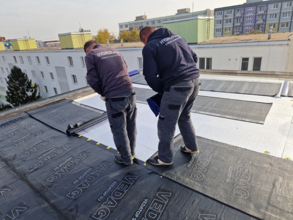 Dva pracovníci firmy Hydros Bánovce v tmavom pracovnom oblečení realizujú hydroizoláciu plochej strechy v meste. Na biely podklad ukladajú a vyrovnávajú čierne asfaltové pásy značky Vedag, pričom v pozadí vidno mestskú zástavbu s bytovými domami.