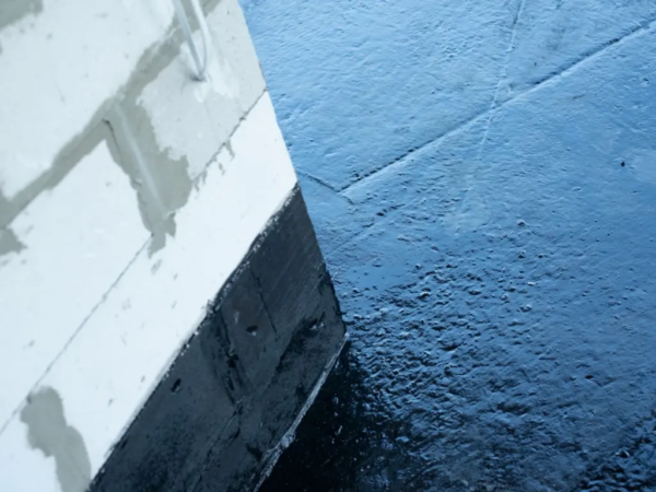 Detailný záber zvrchu na rohovú stenu z pórobetónových tvárnic (ytong), ktorej spodná časť je ošetrená hrubou, lesklou čiernou hydroizolačnou vrstvou, siahajúcou aj na prilahlú vodorovnú podlahovú plochu s nerovnomernou textúrou.