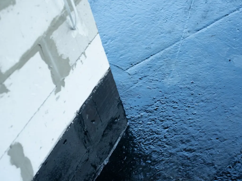 Detailný záber zvrchu na rohovú stenu z pórobetónových tvárnic (ytong), ktorej spodná časť je ošetrená hrubou, lesklou čiernou hydroizolačnou vrstvou, siahajúcou aj na prilahlú vodorovnú podlahovú plochu s nerovnomernou textúrou.